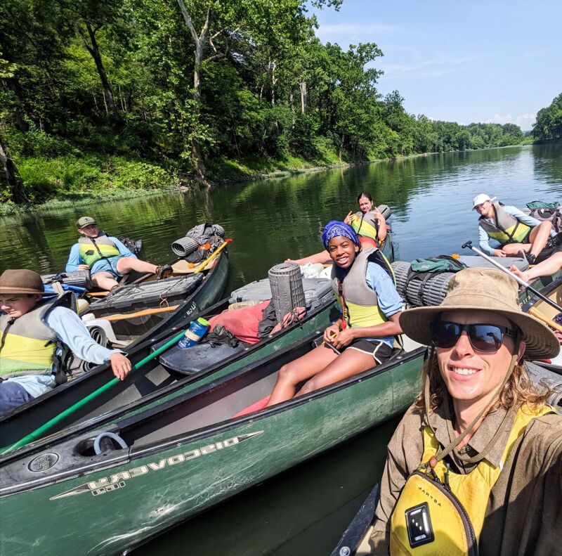 The image shows a group of people canoeing on a river. They are in several canoes, paddling through calm water surrounded by lush green trees. The sky is partly cloudy. Everyone is wearing life vests. The person in the foreground is taking a selfie, wearing a hat and sunglasses.